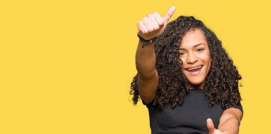 Kıvırcık saçlı genç güzel kadın elle olumlu jest yapıyor onaylayan, başparmak kadar gülümseyerek ve başarı için mutlu. Kameraya bakmak, kazanan jest.