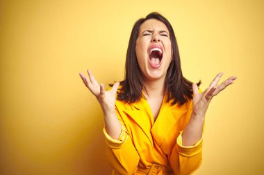Genç güzel esmer kadın sarı izole arka plan deli ve deli bağırarak ve agresif ifade ve kollar kaldırdı ile bağırıyor üzerinde zarif bir elbise giyiyor. Hayal kırıklığı kavramı.