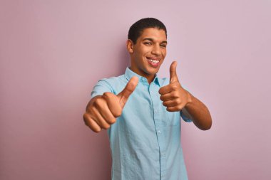 İzole pembe arka plan üzerinde duran mavi gömlek giyen genç yakışıklı Arap adam el ile olumlu jest yapıyor onaylayan, gülümseyerek ve başarı için mutlu başparmak. Kazanan jest.