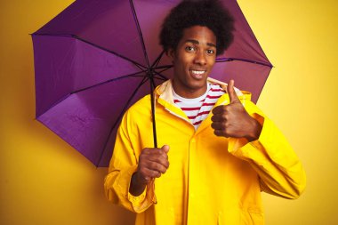Afro amerikan adam yağmur ceket ve şemsiye giyen izole sarı arka plan üzerinde büyük bir gülümseme ok işareti yapıyor mutlu ayakta, parmakları ile başparmak, mükemmel işareti