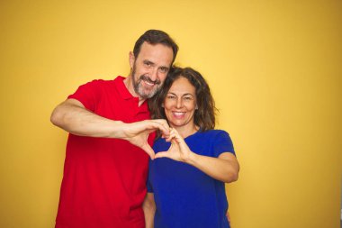 Güzel orta yaş çift birlikte izole sarı arka plan üzerinde sevgi kalp sembolü ve elleriyle şekil gösteren gülümseyerek ayakta. Romantik kavram.