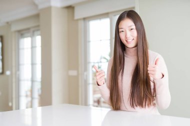 Güzel Asyalı kadın beyaz masa başarı işareti üzerinde rahat kazak giyen el ile olumlu jest yapıyor, gülümseyen ve mutlu başparmak kadar. Neşeli ifade ile kameraya bakmak, kazanan jest.