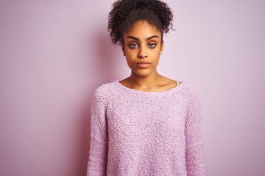 İzole pembe arka plan üzerinde duran kış kazak giyen genç afro-amerikan kadın yüzünde ciddi ifade ile rahat. Kameraya basit ve doğal bir bakış.