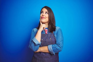 Young beautiful business woman wearing store uniform apron over blue isolated background with hand on chin thinking about question, pensive expression. Smiling with thoughtful face. Doubt concept. clipart