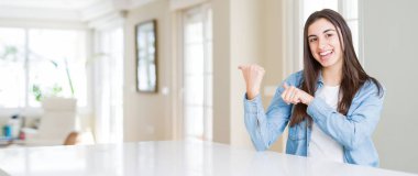 Evde beyaz masada oturan güzel genç kadının geniş açılı resmi El ve başparmakyukarı arkaya işaret, kendinden emin gülümseyen