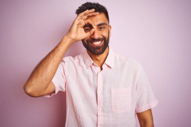 Izole pembe arka plan üzerinde duran rahat gömlek giyen genç hintli adam el gülümseyerek ok jest yapıyor, mutlu yüz ile parmakları ile bakarak göz.