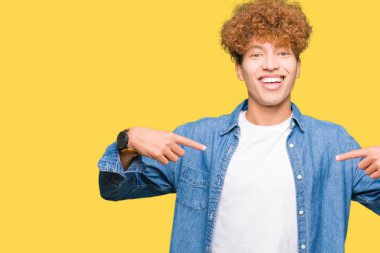 Afro saçlı, kot ceket giyen genç yakışıklı adam yüzünde gülümseme ile kendinden emin görünümlü, parmakları gururlu ve mutlu kendini işaret.