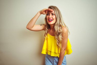 Beyaz izole arka plan üzerinde duran sarı t-shirt giyen genç güzel kadın çok mutlu ve başının üzerinde el ile uzak bakarak gülümsüyor. Arama konsepti.