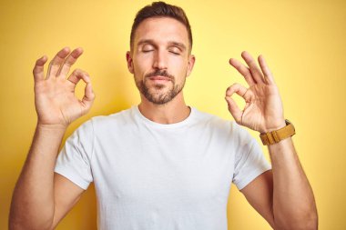 Genç yakışıklı adam sarı izole arka plan üzerinde rahat beyaz t-shirt giyen rahat ve gözleri parmakları ile meditasyon jest yapıyor kapalı gülümseyerek. Yoga konsepti.