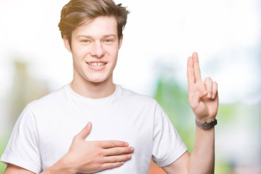 Casual beyaz t-shirt üzerinde izole giyen genç yakışıklı adam arka plan Swearing göğüs ve parmaklar, el ile bir sadakat sözü yemin yapma