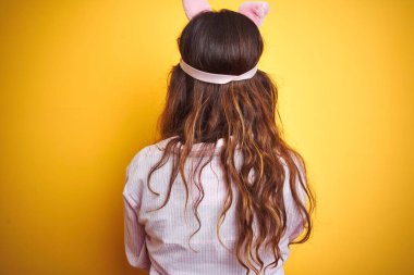 Pijama ve uyku maskesi giyen genç kadın sarı izole arka plan üzerinde duran geriye doğru çapraz kolları ile uzağa bakarak ayakta