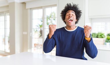 Evde oturan rahat kazak giyen genç Afro-Amerikan adam başarı için heyecanlı kolları kaldırdı ve gözleri kapalı zafer gülümseyerek kutluyor. Kazanan kavramı.