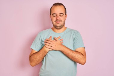 Izole pembe arka plan üzerinde duran mavi rahat t-shirt giyen genç adam kapalı gözleri ve yüzünde minnettar jest ile göğüs üzerinde gülümseyerek. Sağlık kavramı.