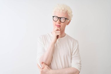 Genç albino sarışın adam t-shirt ve gözlük giyen çene üzerinde el ile izole beyaz arka plan üzerinde duran soru, dalgın ifade düşünme. Düşünceli bir yüzle gülümsüyor. Şüphe kavramı.