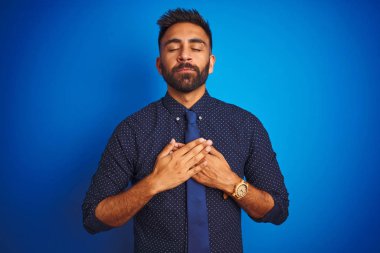 Zarif gömlek ve kravat giyen genç Hintli işadamı kapalı gözleri ve yüzünde minnettar jest ile göğüs üzerinde gülümseyerek izole mavi arka plan üzerinde ayakta. Sağlık kavramı.