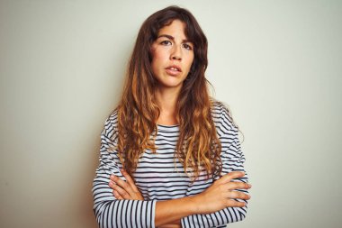 Beyaz izole arka plan şüpheci ve sinir üzerinde çizgili t-shirt giyen genç güzel kadın, çapraz kolları ile yüzünde ifade onaylamayan. Negatif kişi.