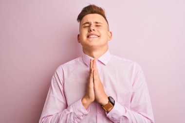 İzole edilmiş pembe arka plan üzerinde duran zarif gömlek giyen genç yakışıklı işadamı çok duygusal ve endişeli yüzünde umut ifadesi ile birlikte elleri ile dua. Af dilemek istiyorum. Din kavramı.