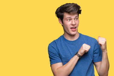 İzole arka plan üzerinde mavi t-shirt giyen genç yakışıklı adam el ve başparmak kadar arka arkasında Işaret, gülümseyen emin