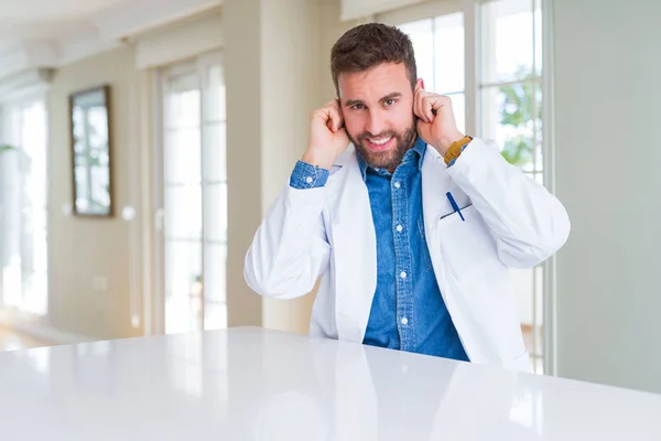 Handsome doctor man wearing medical coat at the clinic covering ears with fingers with annoyed expression for the noise of loud music. Deaf concept.