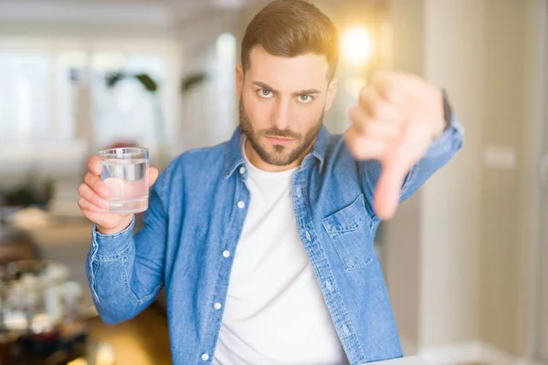 年轻的帅哥在家里喝着一杯水 生气的脸 消极的标志显示不喜欢用拇指向下 拒绝概念 — 图库照片
