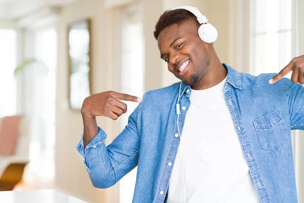 アフリカ系アメリカ人の顔に笑顔と自信を持って 誇りと幸せな指で自分を指して音楽を聴くヘッドフォンを身に着けています — ストック写真