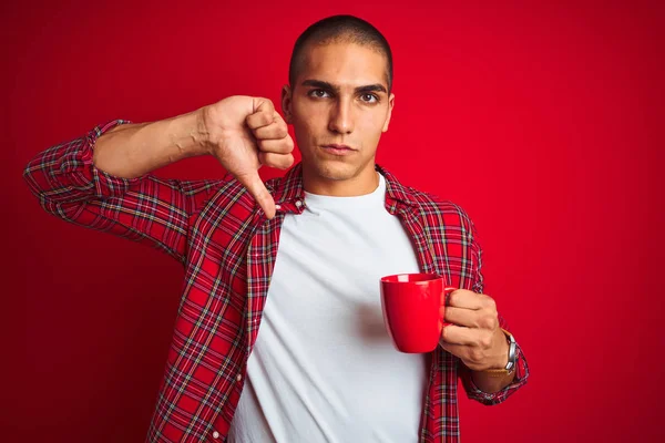 Young handsome man wearing shirt drinking a cup of coffee over red isolated background with angry face, negative sign showing dislike with thumbs down, rejection concept