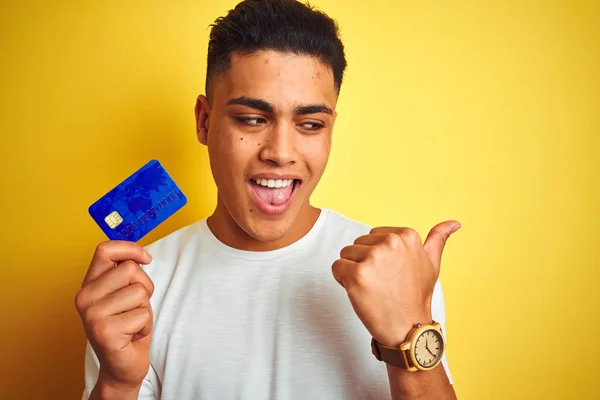 年轻的巴西客户男子拿着信用卡站在孤立的黄色背景指向和显示与大拇指的一边与快乐的脸微笑 — 图库照片