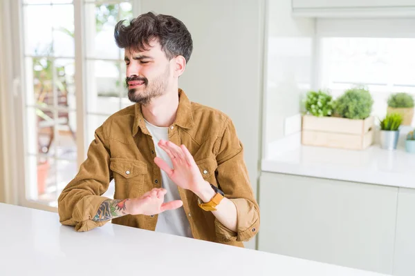 Young man wearing casual jacket sitting on white table disgusted expression, displeased and fearful doing disgust face because aversion reaction. With hands raised. Annoying concept.