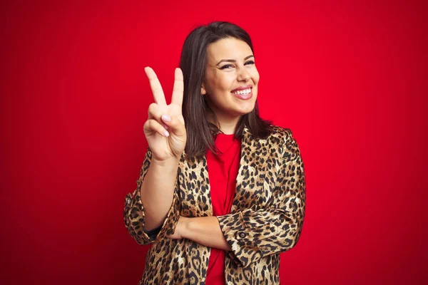 Young beautiful brunette woman wearing a leopard jacket over red isolated background smiling with happy face winking at the camera doing victory sign. Number two.