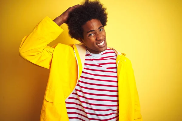 African american man with afro hair wearing rain coat standing over isolated yellow background confuse and wonder about question. Uncertain with doubt, thinking with hand on head. Pensive concept.