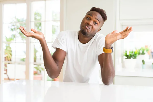 英俊的非洲裔美国男子在家里的白色桌子上 不知所措 用手臂和双手举起糊涂的表情 怀疑概念 — 图库照片