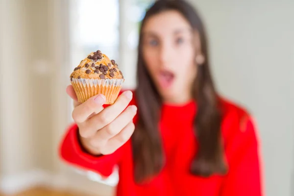 チョコレートチップマフィンを食べる美しい若い女性は 驚きの顔でショックを受け 恐怖表現で恐怖と興奮 — ストック写真