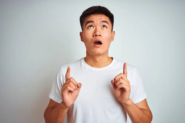 年轻的亚洲中国男子穿着T恤站在孤立的白色背景惊讶和惊讶抬头 用手指指着和抬起手臂 — 图库照片