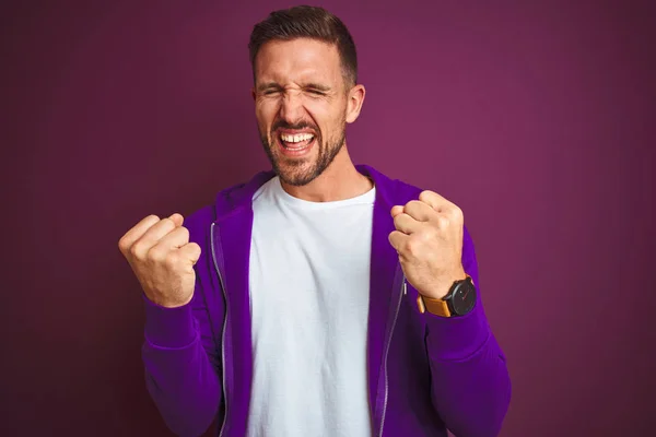 Young fitness man wearing casual sports sweatshirt over purple isolated background very happy and excited doing winner gesture with arms raised, smiling and screaming for success. Celebration concept.