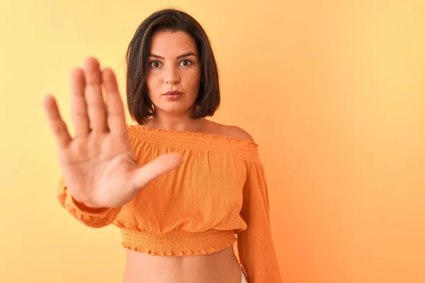 Young beautiful woman wearing casual t-shirt standing over isolated orange background doing stop sing with palm of the hand. Warning expression with negative and serious gesture on the face.