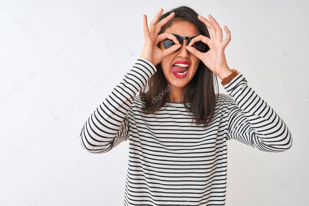 Chinese woman wearing striped t-shirt and sunglasses standing over isolated white background doing ok gesture like binoculars sticking tongue out, eyes looking through fingers. Crazy expression.