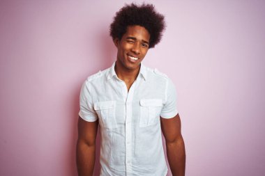 Beyaz gömlek giyen afro saçlı genç amerikalı adam izole pembe arka plan üzerinde duran seksi ifade, neşeli ve mutlu yüz ile kameraya bakarak göz kırpıyor.
