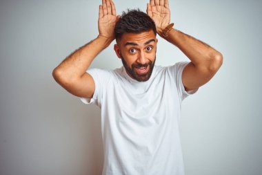 İzole beyaz arka plan üzerinde duran t-shirt giyen genç hintli adam alaycı ve şüpheci görünümlü avuç içi ile tavşan kulakları jest yapıyor. Paskalya tavşanı kavramı.