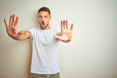 Genç yakışıklı adam izole arka plan üzerinde rahat beyaz t-shirt giyen avuç içi, kızgın ve hayal kırıklığı ifade ile stop jest yapıyor