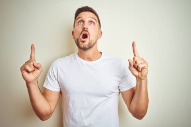 Yakışıklı delikanlı izole arka plan üzerinde rahat beyaz t-shirt giyen hayrete ve yukarıya bakıyor ve parmakları ile işaret ve silah kaldırdı şaşırttı.