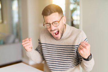 Evde gözlük takan genç yakışıklı adam çok mutlu ve heyecanlı kollarını kaldırdı, gülümseyerek ve başarı için çığlık ile kazanan jest yapıyor. Kutlama konsepti.