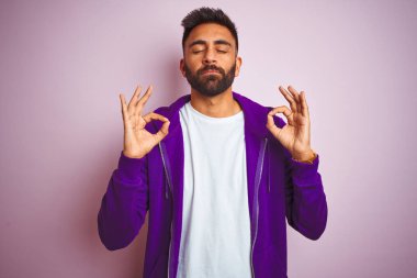 İzole pembe arka plan üzerinde duran mor sweatshirt giyen genç hintli adam rahatlayın ve parmakları ile meditasyon jest yapıyor kapalı gözleri ile gülümseyerek. Yoga konsepti.