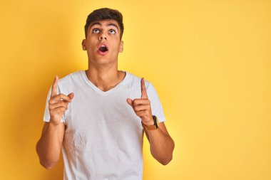 İzole sarı arka plan üzerinde duran beyaz t-shirt giyen genç hintli adam şaşırttı ve yukarı bakarak ve parmakları ile işaret ve kolları kaldırdı şaşırttı.
