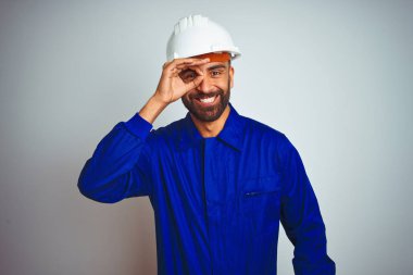 Yakışıklı hint işçi adam izole beyaz arka plan üzerinde üniforma ve kask giyen el gülümseyerek ok jest yapıyor, mutlu yüz ile parmaklardan bakan göz.