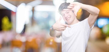 Elle yapma çerçeve ve parmakları ile mutlu yüz gülümseyen izole arka plan üzerinde rahat beyaz t-shirt giyen genç yakışıklı adam. Yaratıcılık ve Fotoğrafçılık kavramı.