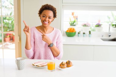 Genç afro-amerikan kadın evde sabah breaksfast yeme gülümseyerek ve yan iki el ve parmakları ile işaret kameraya bakarak.