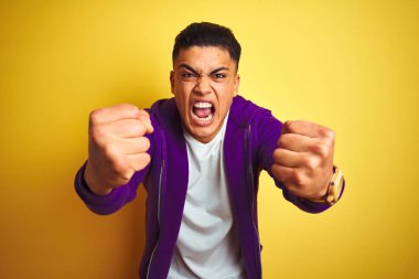 Mor sweatshirt giyen genç brezilyalı adam izole sarı arka plan üzerinde duran kızgın ve öfkeli öfke ile bağırırken sinirli ve öfkeli yumruklar yükselterek. Öfke ve agresif kavram.