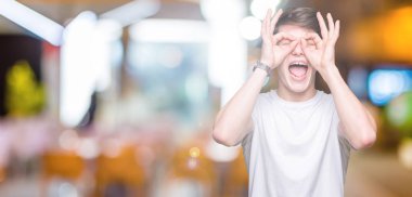 Dilini, parmaklarının arasından baktığını yapışmasını dürbün gibi ok jest yapıyor izole arka plan üzerinde rahat beyaz t-shirt giyen genç yakışıklı adam. Deli ifade.