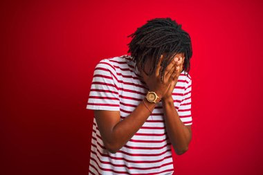 Dreadlocks çizgili t-shirt giyen genç afro adam üzgün ifade elleri ile yüz kapsayan izole kırmızı arka plan üzerinde duran ağlarken. Depresyon kavramı.