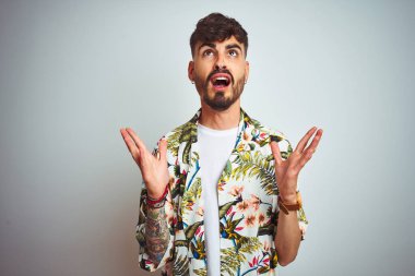 Tatilde dövmeli adam yazlık gömlek giyen izole beyaz arka plan üzerinde duran deli ve deli bağırarak ve agresif ifade ve kollar kaldırdı ile bağırıyor. Hayal kırıklığı kavramı.